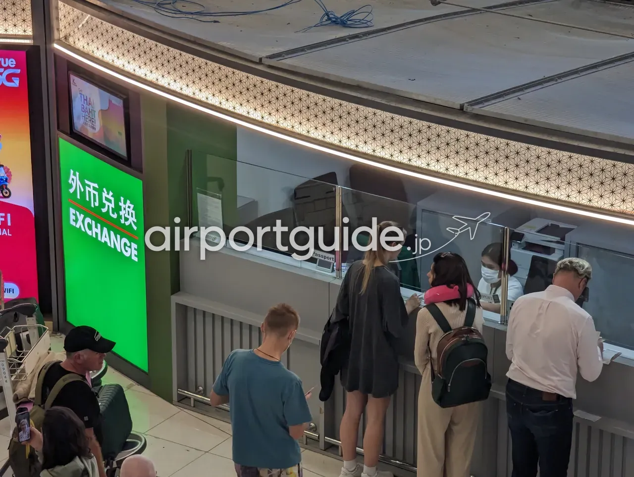 スワンナプーム空港の外貨両替カウンター。緑地に「外币兑换 EXCHANGE」、モニターに『THAI BAHT HERE!』の表示。カウンターにはPassportトレイがあり、旅行者が両替の手続きをしている。