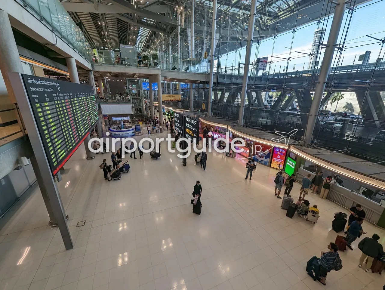 スワンナプーム空港の国際線到着ホールを上階から俯瞰。左に大型の「International Arrivals Door B」到着便案内板、中央にAOTリムジンのカウンター、右手にガラス壁と待合、スーツケースを引く旅客が行き交う。