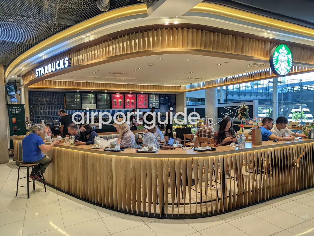 スワンナプーム空港内のスターバックス。木のルーバーで縁取られたカウンターと客席、「STARBUCKS」の看板とセイレーンのロゴ、ガラス越しに高架のレールリンクが見える。