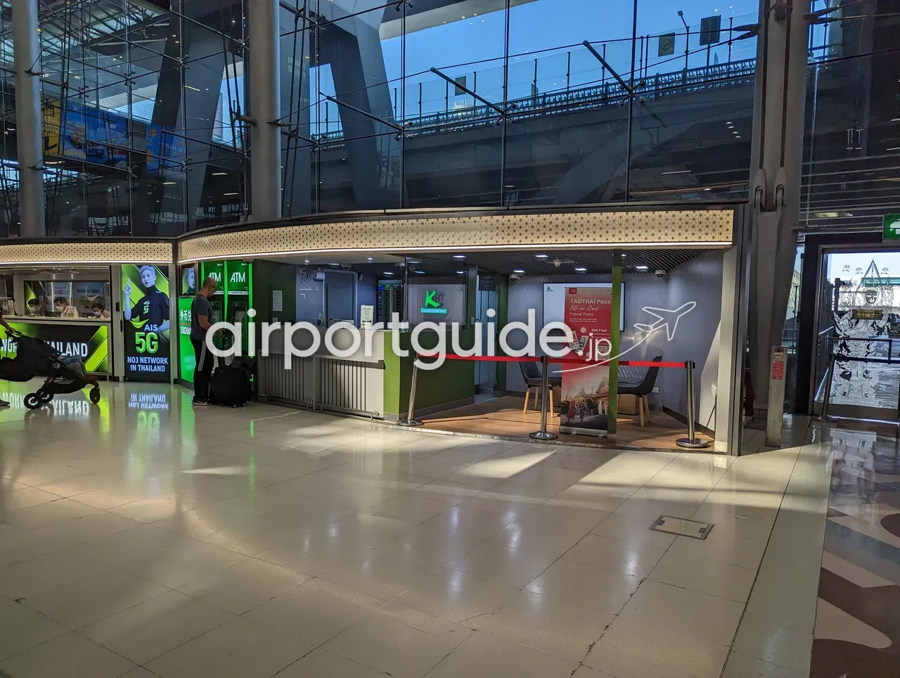 スワンナプーム空港の到着階のサービスカウンター。「ATM」機と緑の「K KASIKORNTHAI」ブース、「TAGTHAI Pass All-in-One Travel Pass」の案内、AIS 5Gの広告、ガラス壁の外に高架のレールリンク。