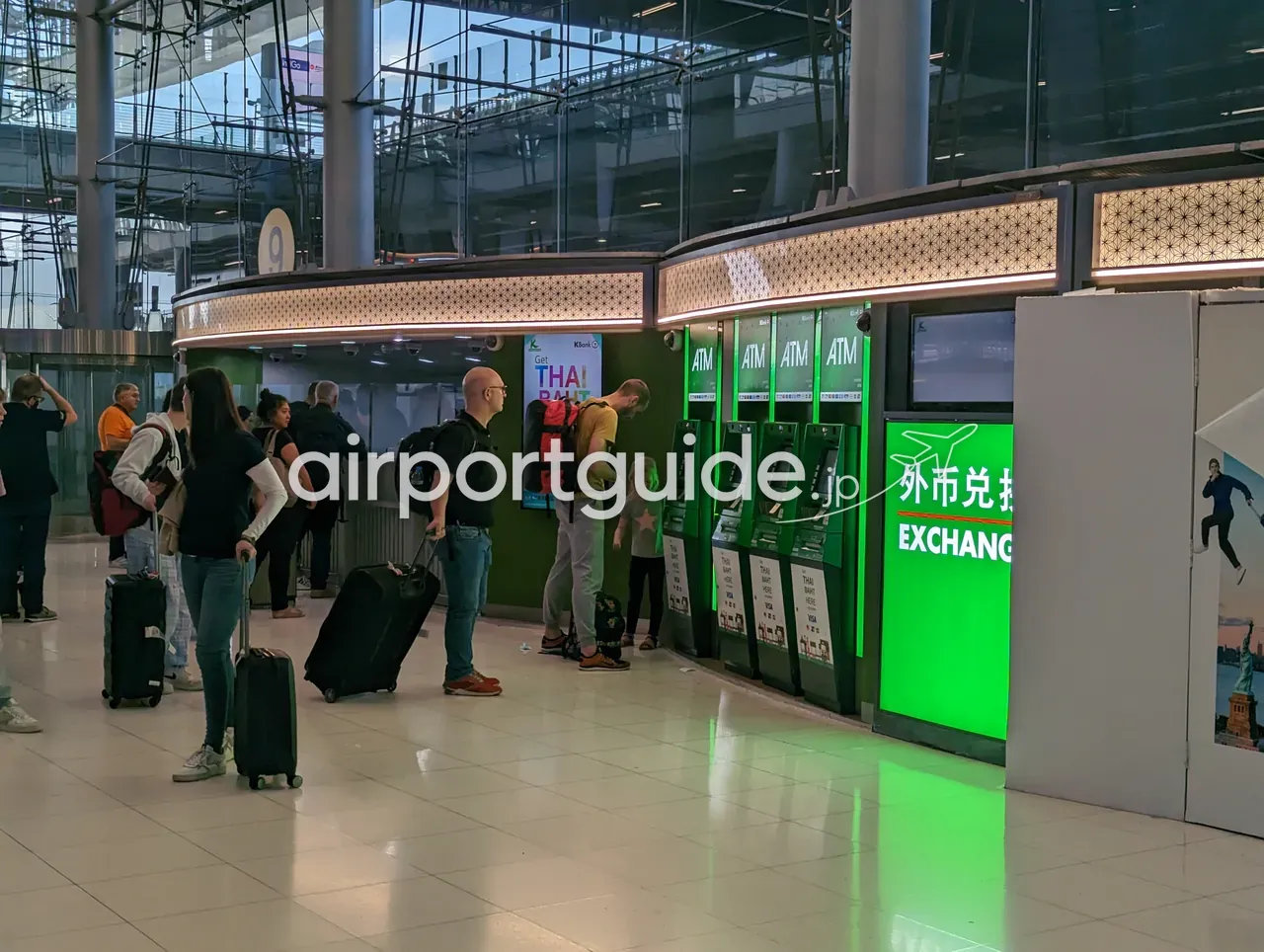 スワンナプーム空港の到着階のATMコーナーと外貨両替ブース。緑の「EXCHANGE（外币兑换）」看板と4台並んだ「ATM」、「Get THAI BAHT」の広告、スーツケースを持つ旅行者が両替を待つ。