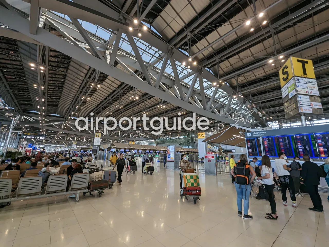 スワンナプーム空港の出発チェックイン階。黄色い「S／T ROW S-T」のサインに航空会社ロゴ（大韓航空・エバー航空ほか）が並び、右手に大型の出発便案内板（Departures）、うねる鉄骨大屋根が広がる。