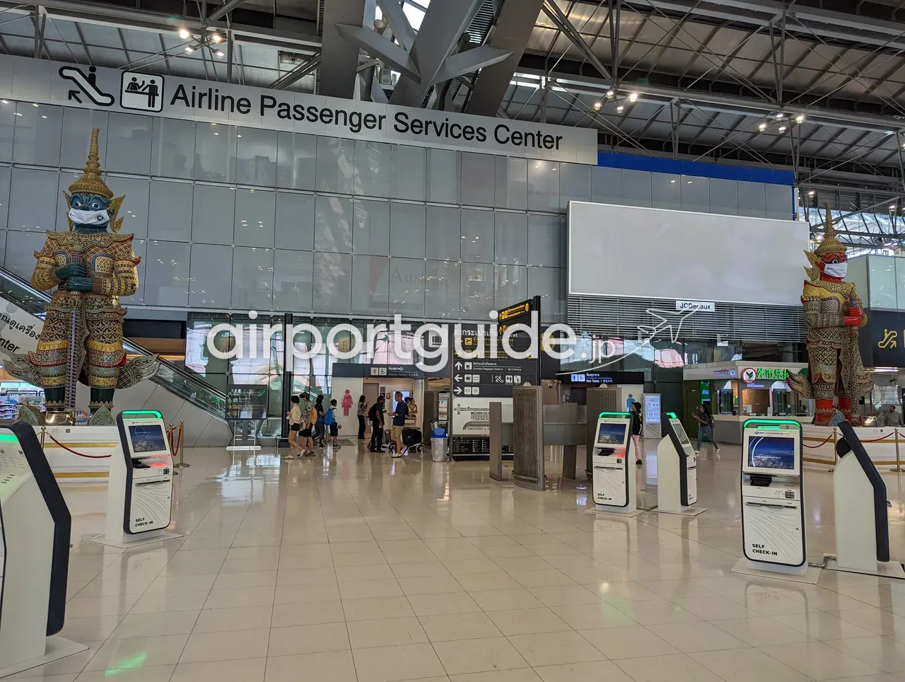 スワンナプーム空港の出発チェックイン階。「Airline Passenger Services Center」の案内サインの下、左右にマスクを着けた2体の巨大な金色のヤック(守護神)像が立ち、手前にセルフチェックイン機、右手に両替(EXCHANGE)ブースが並ぶ。