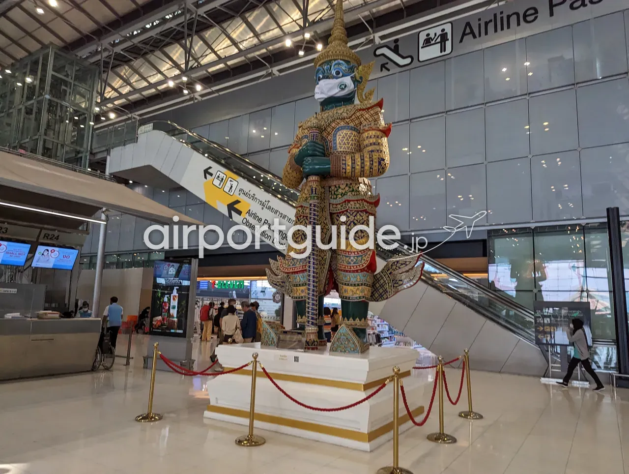 スワンナプーム空港のチェックイン階に立つ、マスクを着けた巨大な金色のヤック（守護神）像。白い台座と赤金のロープ柵、頭上に「Airline Passenger Services Center／Observation Deck」の案内サイン。