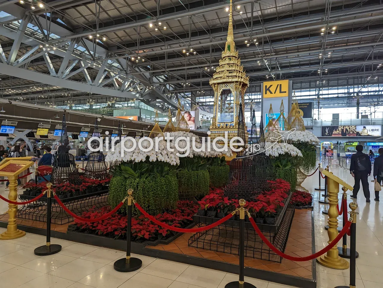 スワンナプーム空港の出発チェックインホールに設けられた、タイ様式の金色の楼閣（ブサボック）と白い胡蝶蘭・赤いポインセチアの装飾。奥に黄色い「K／L」の出発案内サイン。