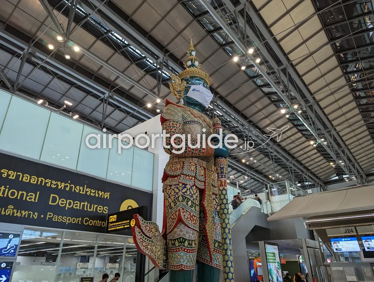 スワンナプーム空港の巨大な金色のヤック（守護神）像を見上げた構図。マスクを着けた像の左に「International Departures・Passport Control（出国審査）」の黒い案内サイン、右奥にゲートJ24。