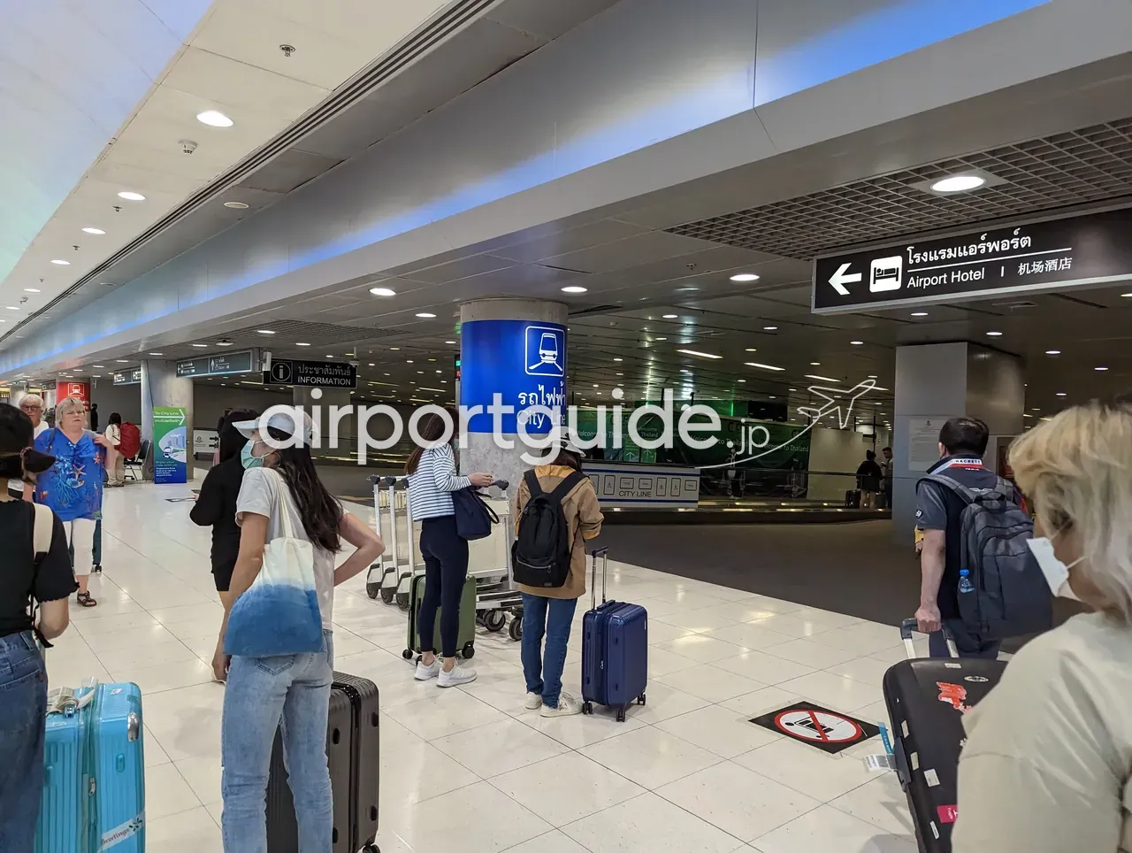 スワンナプーム空港のエアポート・レール・リンク乗り場付近。青い「City Line（รถไฟฟ้า）」の柱サインと「Welcome to Thailand」の緑の壁、頭上に「Airport Hotel（机场酒店）」への矢印案内、スーツケースを持つ旅客が並ぶ。