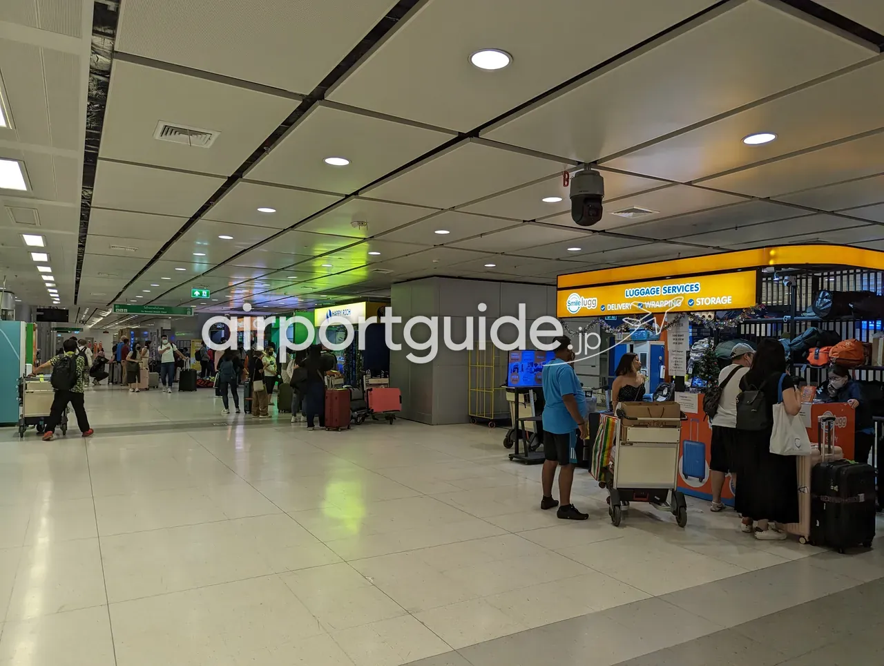 スワンナプーム空港のレールリンク階の通路にある「Smilelugg LUGGAGE SERVICES（DELIVERY・WRAPPING・STORAGE）」の手荷物サービスカウンター。荷物を預ける旅客と、奥に両替ブースの列。