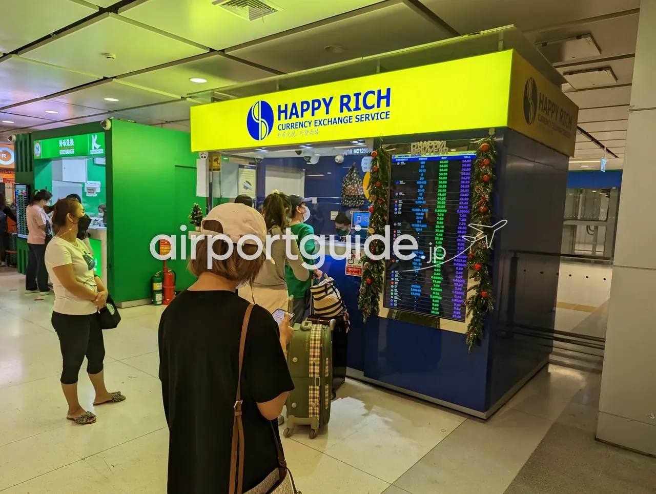 スワンナプーム空港・レールリンク階のHAPPY RICH Currency Exchange Serviceのブース。黄色い看板とBUY/SELLのレート電光板、クリスマスの装飾。旅行者がスーツケースを引いて並んでいる。左隣に緑のカシコン銀行EXCHANGE、右奥にSuperRich。