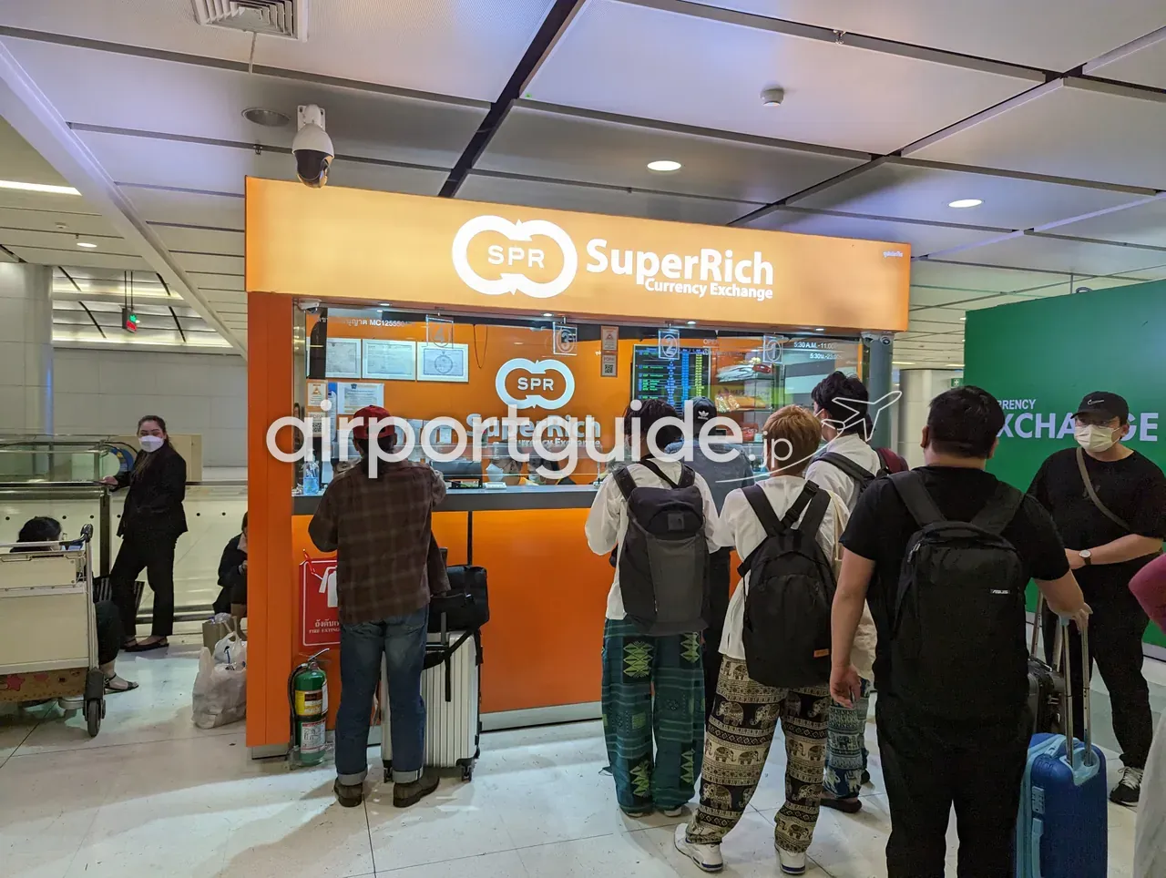 スワンナプーム空港・レールリンク階のSuperRich Currency Exchange両替ブース。オレンジの看板に『SPR SuperRich Currency Exchange』、カウンターにスーツケースの旅行者が列を作る。