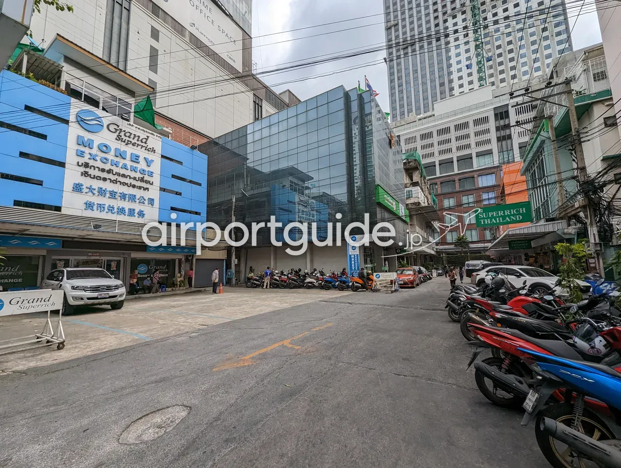 バンコク・プラトゥーナムの両替店街。左に青い『Grand Superrich MONEY EXCHANGE 盛大财宝』のビル、右に緑の『SUPERRICH THAILAND』の看板。手前の道路にバイクが多数駐車している。