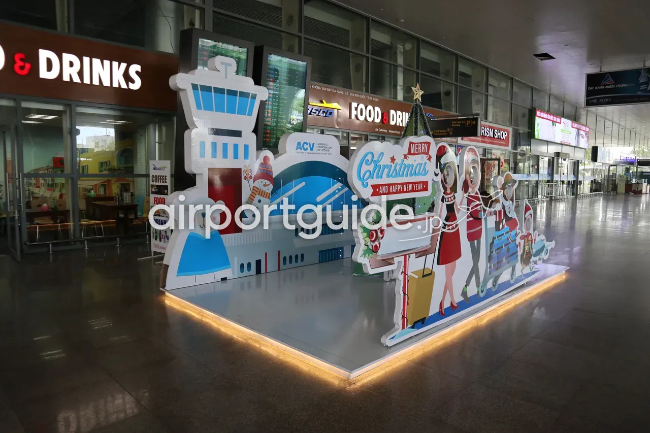 ダナン国際空港ターミナル内のクリスマス装飾。管制塔や飛行機、雪だるま、サンタ、白熊をかたどったパネルと「Merry Christmas and Happy New Year」の飾り、ACVのロゴ。奥に飲食店やショップ、FIDSが並ぶ。