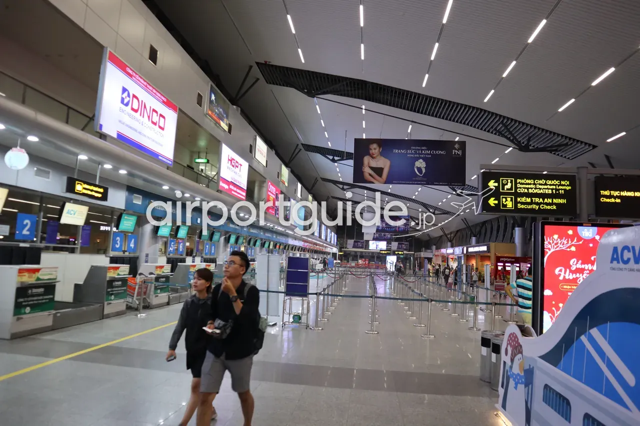 ダナン国際空港・国内線ターミナルの出発ホール。ベトナム航空のチェックインカウンター2〜8番前を旅客が歩き、吊り下げサインに「PHÒNG CHỜ QUỐC NỘI／Domestic Departure Lounge（国内線出発待合）Gates 1-11」「KIỂM TRA AN NINH／Security Check（保安検査）」の案内。ACVの表示。