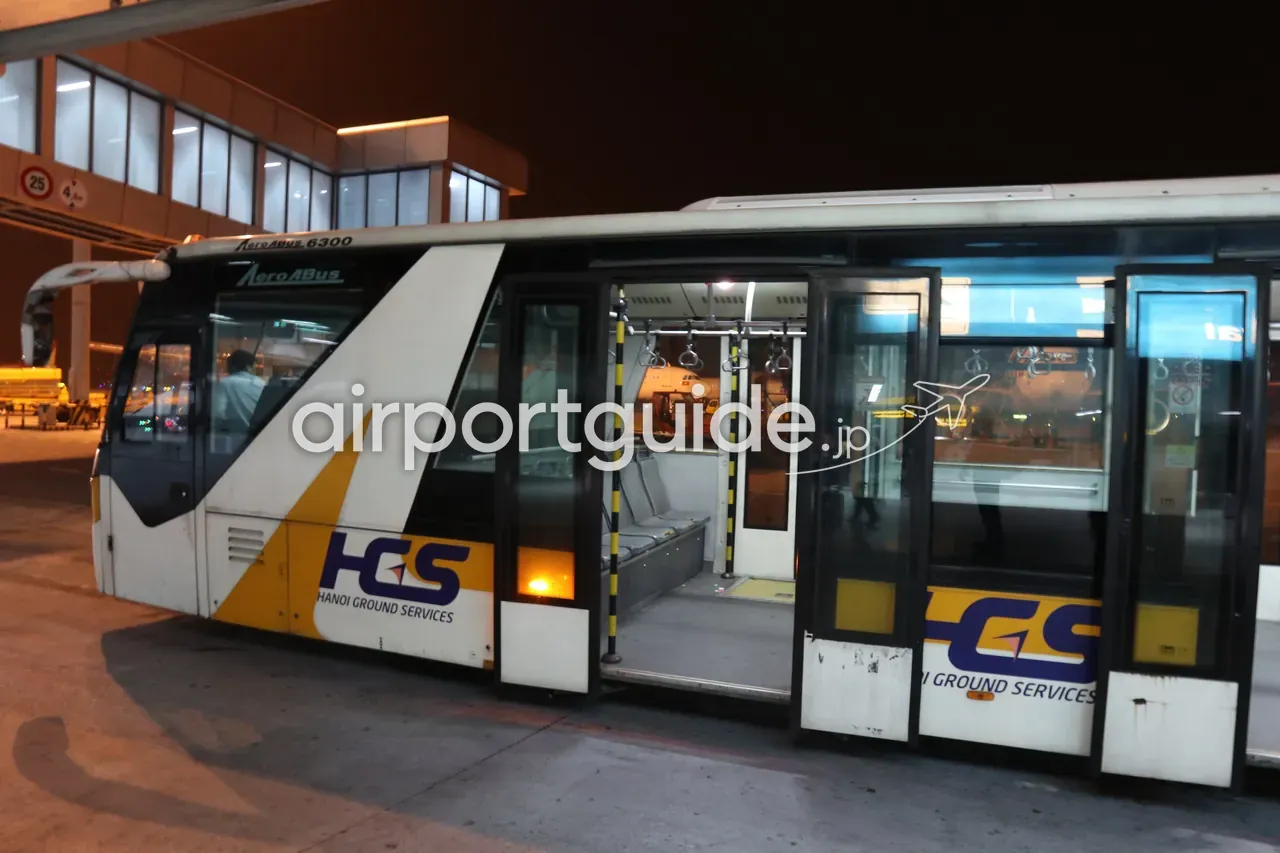 夜のダナン国際空港のエプロンで待機するランプバス（旅客搬送バス）。「HGS／Hanoi Ground Services」のロゴ入り車体、ドアが開き車内の吊り革が見える。搭乗のための地上移動。
