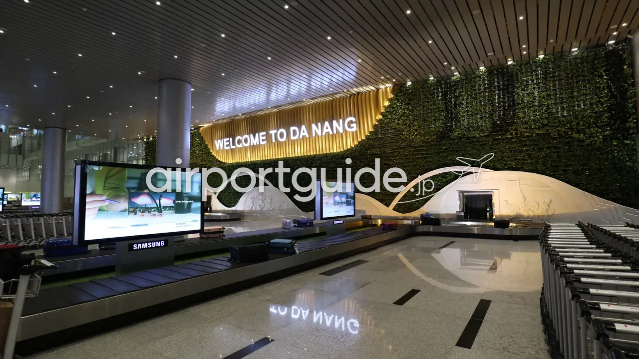 ダナン国際空港・国際線ターミナルの手荷物受取所。「WELCOME TO DA NANG」の光る看板と緑の壁面、波打つ形のターンテーブルにスーツケース、右手に整列した手荷物カート。