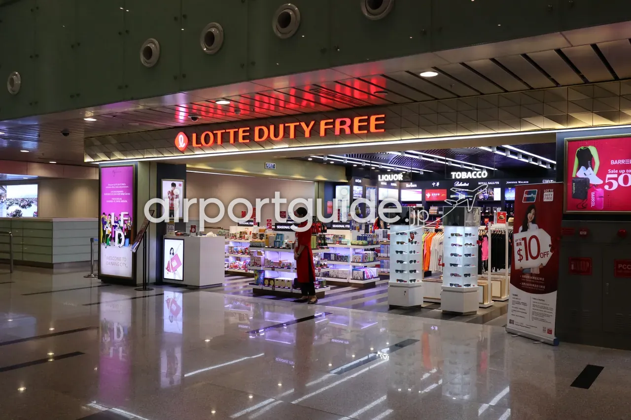 ダナン国際空港・国際線ターミナル制限エリアの「LOTTE DUTY FREE」店頭。LIQUOR（酒）・TOBACCO（たばこ）の棚、サングラスや衣料の陳列、「WELCOME TO DA NANG CITY」の表示、赤いアオザイの店員。