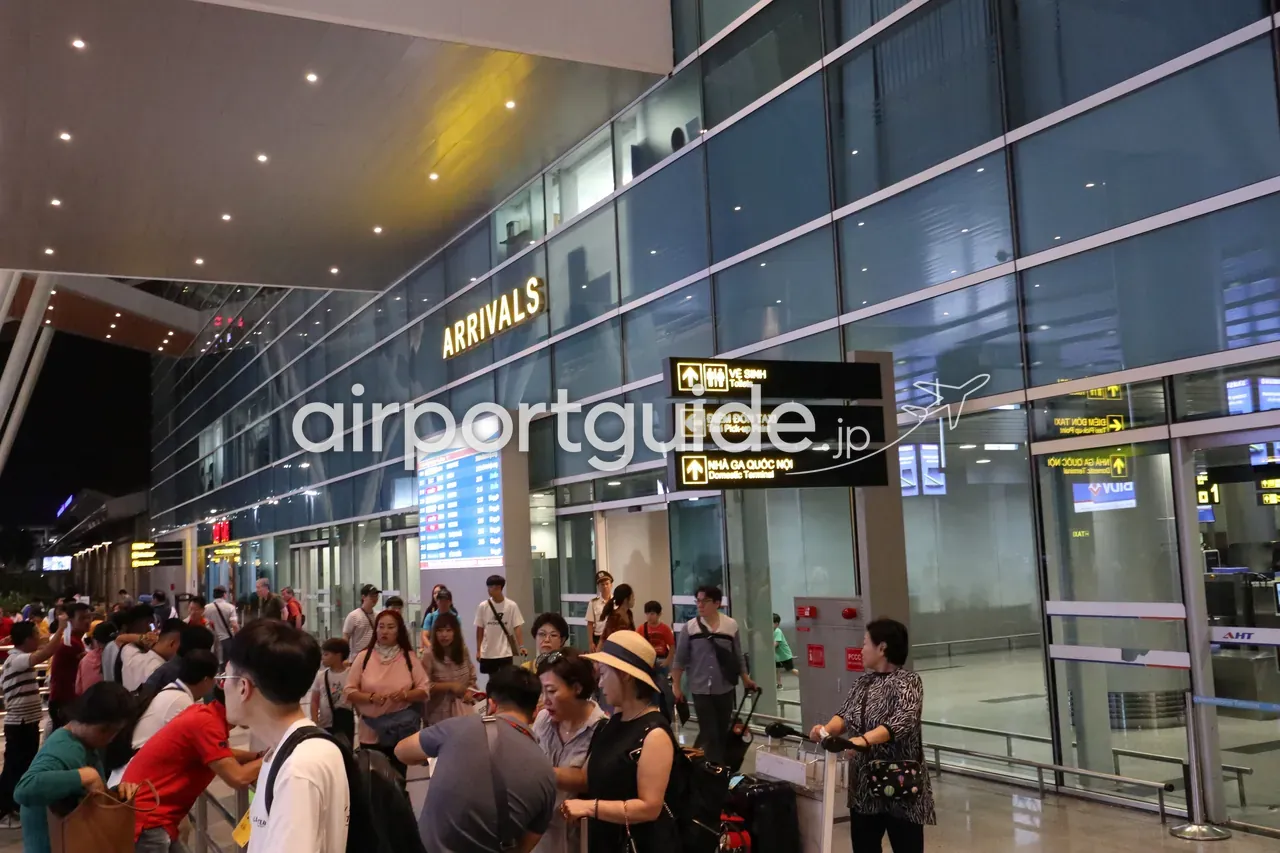 ダナン国際空港・国際線ターミナルの到着口前。金文字の「ARRIVALS」看板とガラス張りのファサード、出迎えや移動の旅客で混雑する。吊り下げ案内には「Taxi Pick-up Point（タクシー乗り場）」「Domestic Terminal（国内線ターミナル）」の矢印。
