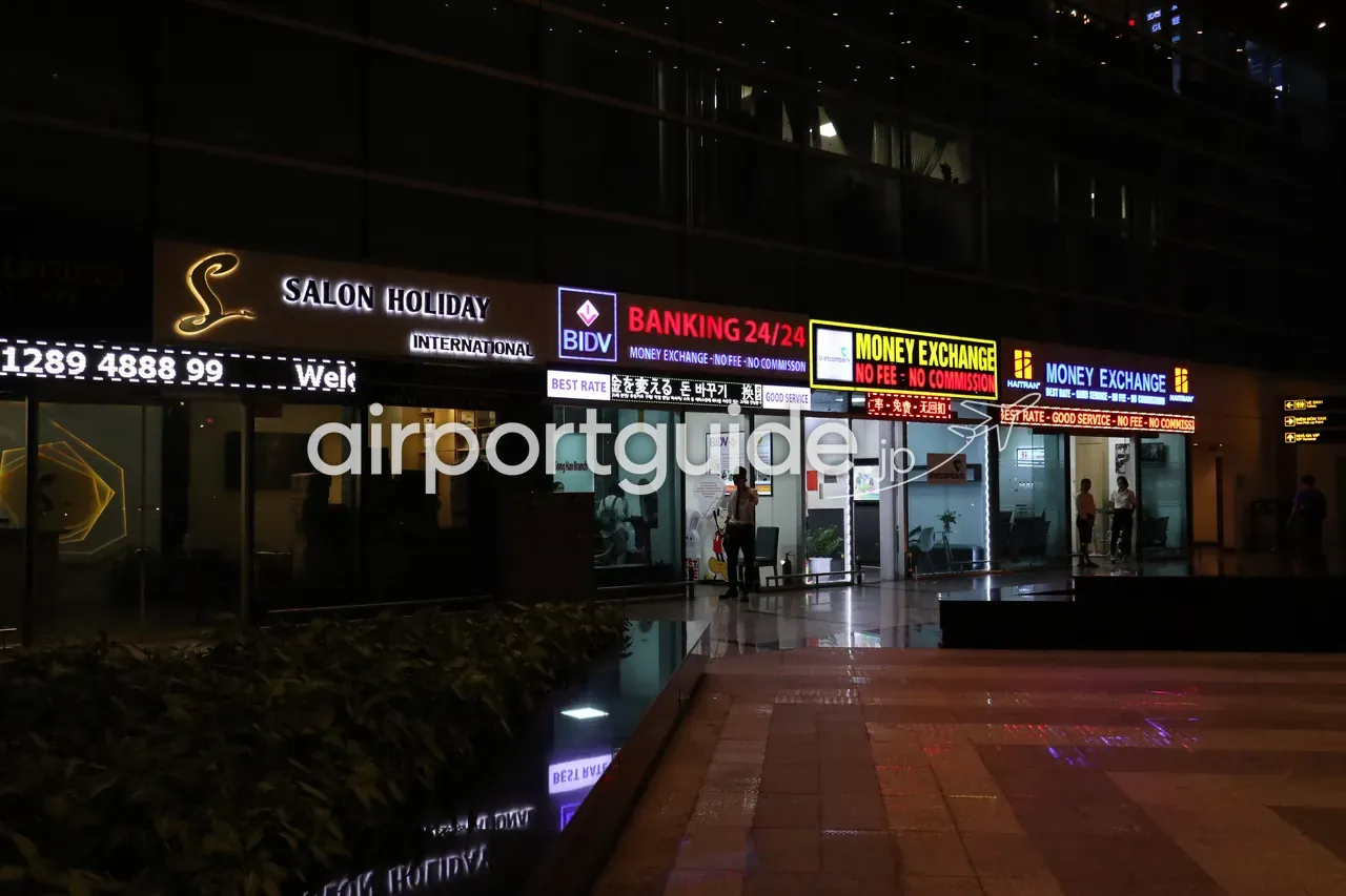 夜のダナン国際空港・国際線ターミナル前の店舗帯。BIDVの「BANKING 24/24」、Vietcombank・Haitranの「MONEY EXCHANGE／NO FEE・NO COMMISSION／BEST RATE」のネオン看板が並び、日本語・韓国語・中国語の表記も。