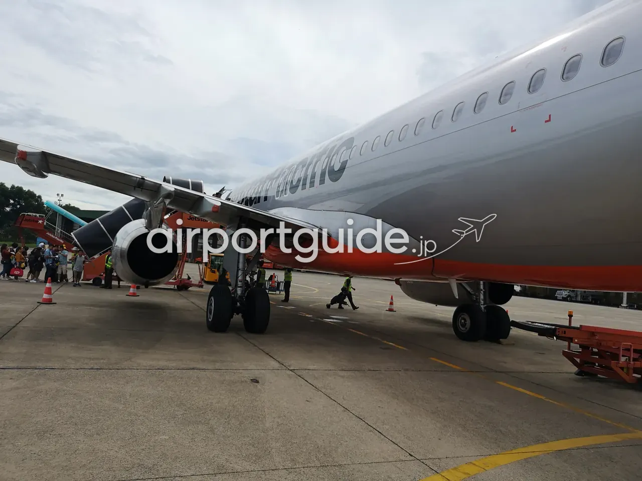 ダナン空港の駐機場（エプロン）。オレンジ塗装のJetstar Pacificの機体にタラップから搭乗する乗客と、蛍光ベストの地上係員、コーン。曇り空。