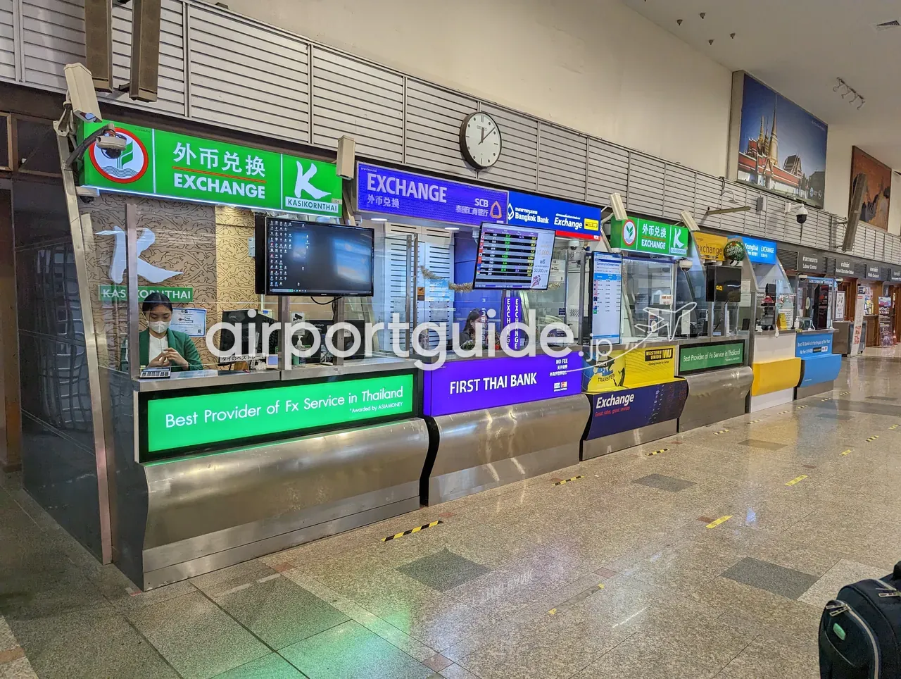 ドンムアン空港の両替所が横一列に並ぶ通路。緑の「KASIKORNTHAI EXCHANGE（外币兑换）」、青の「SCB」、「Bangkok Bank」、紫の「FIRST THAI BANK」の両替カウンターとレート表示板、壁に寺院の写真パネルが掛かる。