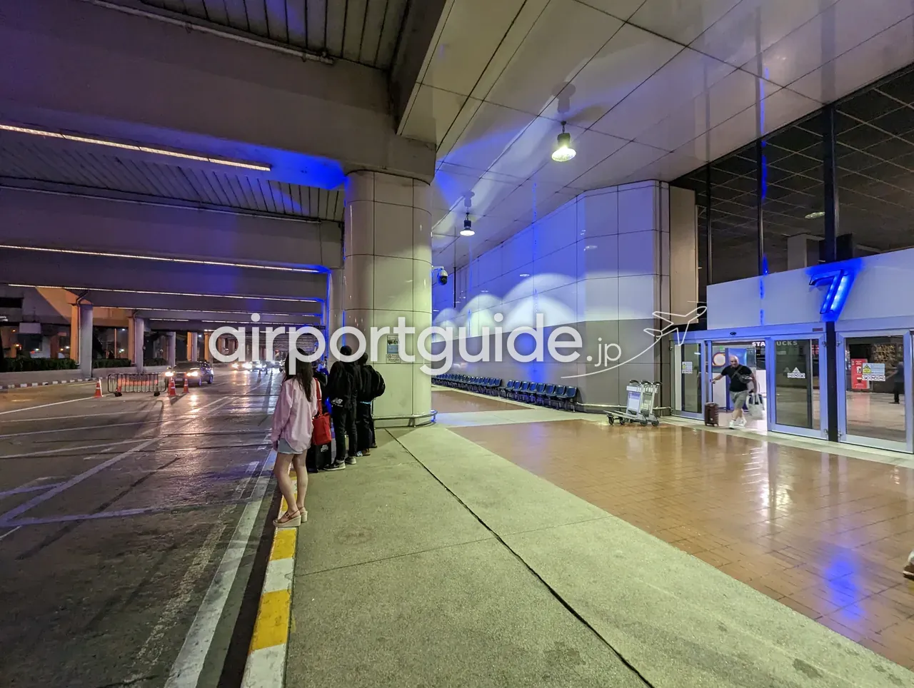 夜のドンムアン空港ターミナル前の車寄せ。青いライトに照らされた歩道で送迎を待つ人々、手荷物カート、奥にスターバックスの入口が見える。