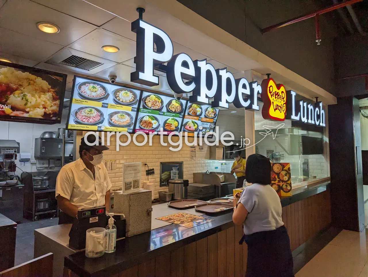 ドンムアン空港内の「Pepper Lunch」の店舗カウンター。黒地に白い「Pepper Lunch」のロゴ看板と、ポークやチキン、サーモンのペッパーライスの写真メニュー（150〜255バーツ）、マスク姿の店員と客。