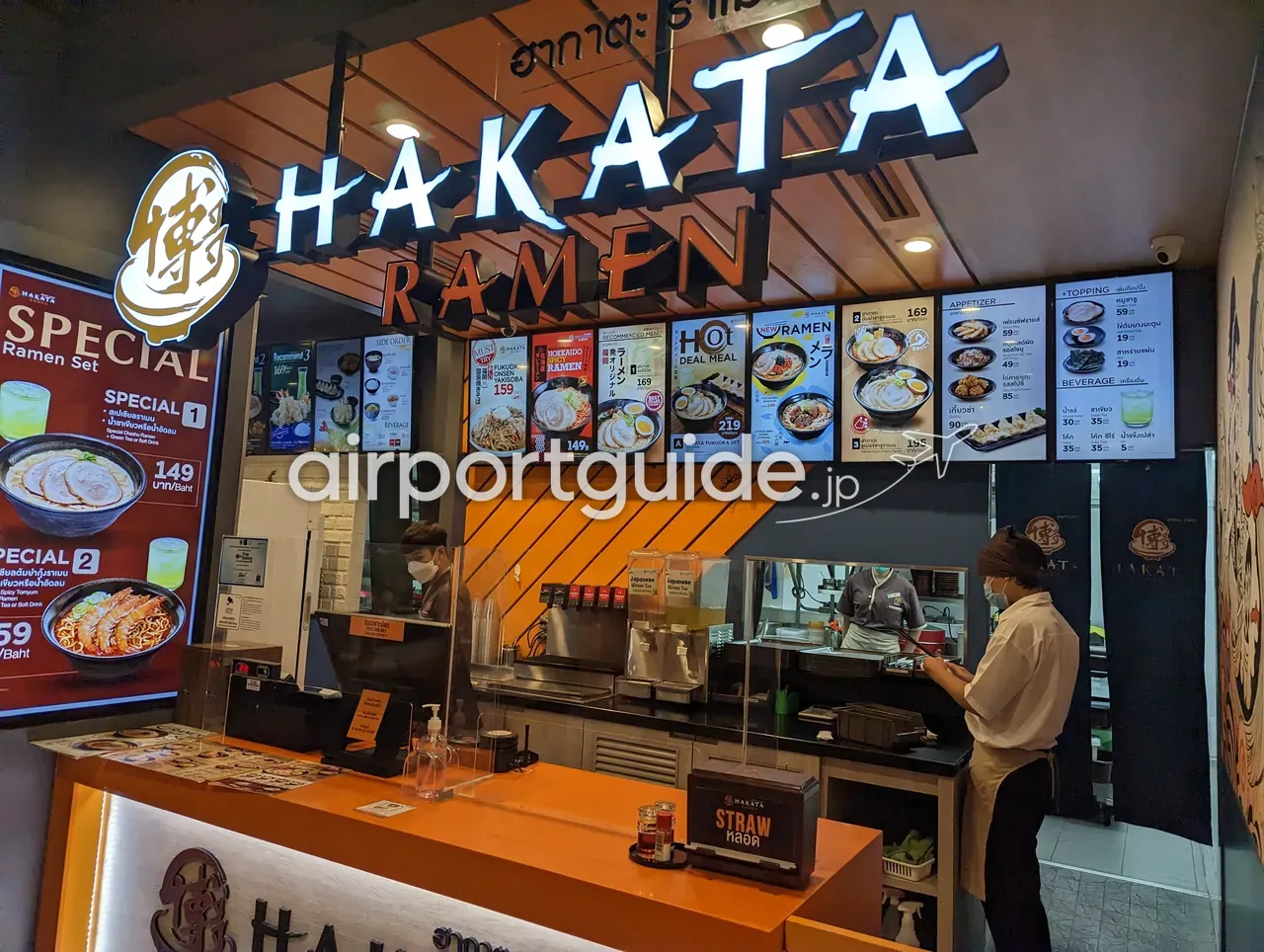 ドンムアン空港内のラーメン店「HAKATA RAMEN（ฮากาตะ ราเมน）」のカウンター。オレンジと白の看板、スペシャルラーメンセットや各種ラーメンの写真メニューが並び、店員が調理している。