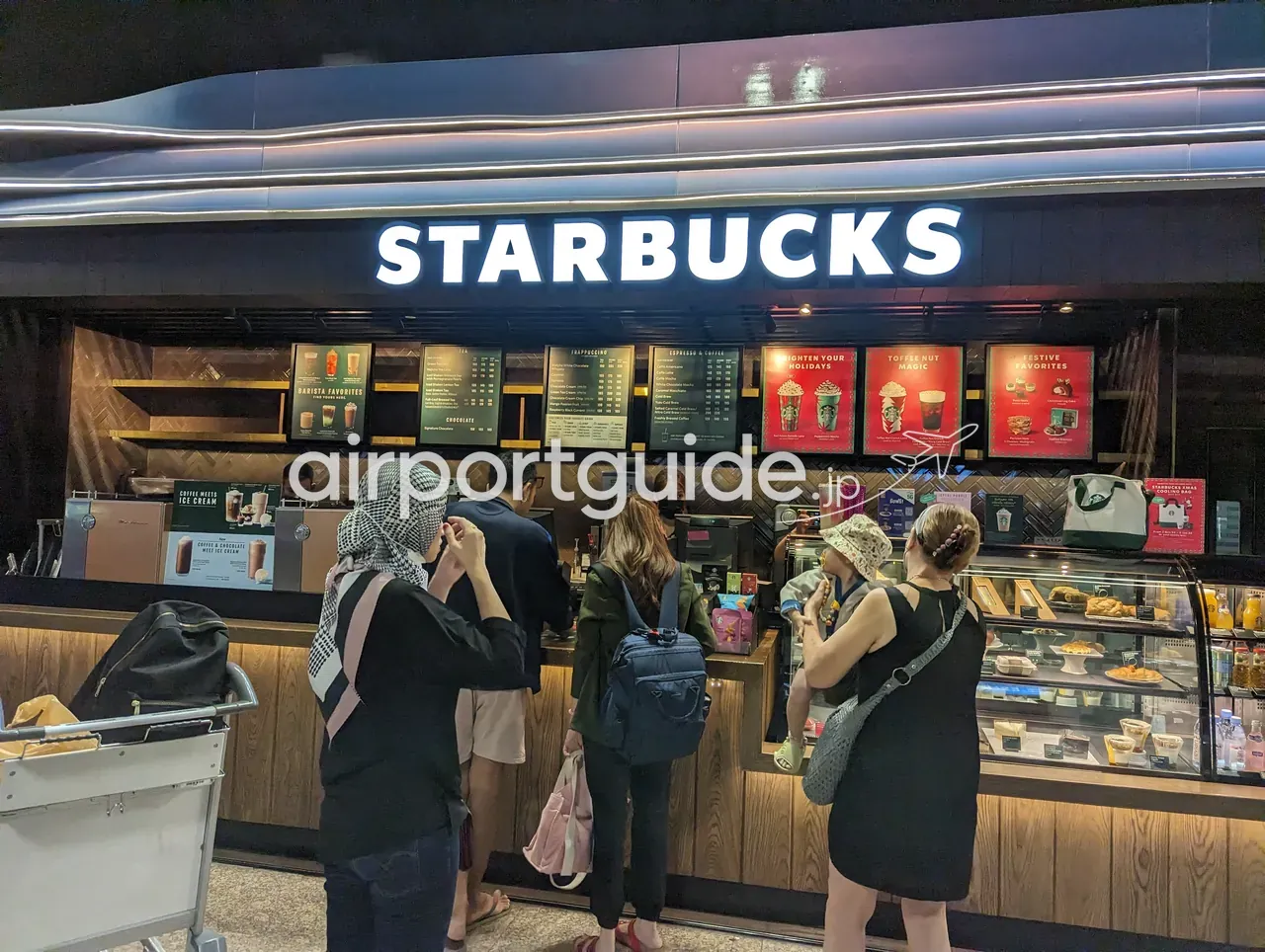 ドンムアン空港内のスターバックス。白い「STARBUCKS」の看板の下にメニューボードとショーケース、木目のカウンター、コーヒーを買い求める旅客の列。