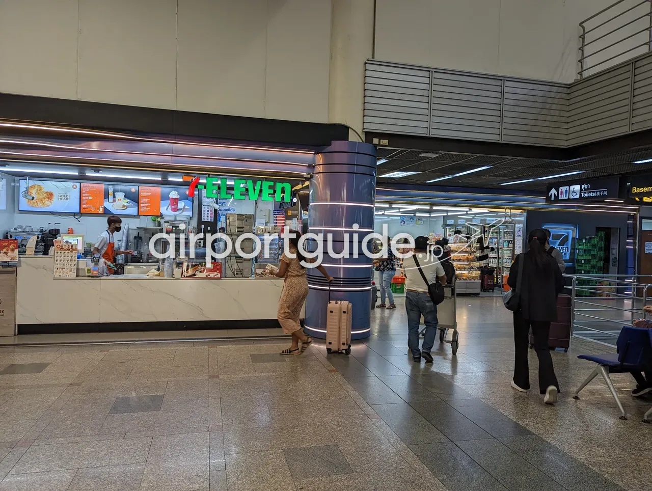 ドンムアン空港内のセブンイレブン。緑の「7-ELEVEN」ロゴのカウンターとコーヒーカウンター、飲料棚が並び、スーツケースを引く旅客が店の前を通る。