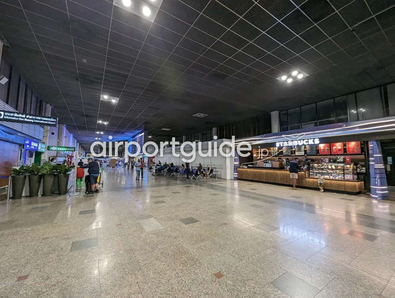 ドンムアン空港のコンコース。「Information Don Muang International Airport」の案内サインと両替(EXCHANGE)ブース、中央のフライト案内板、右手にスターバックスのカウンターが並び、御影石の床を旅客が行き交う。