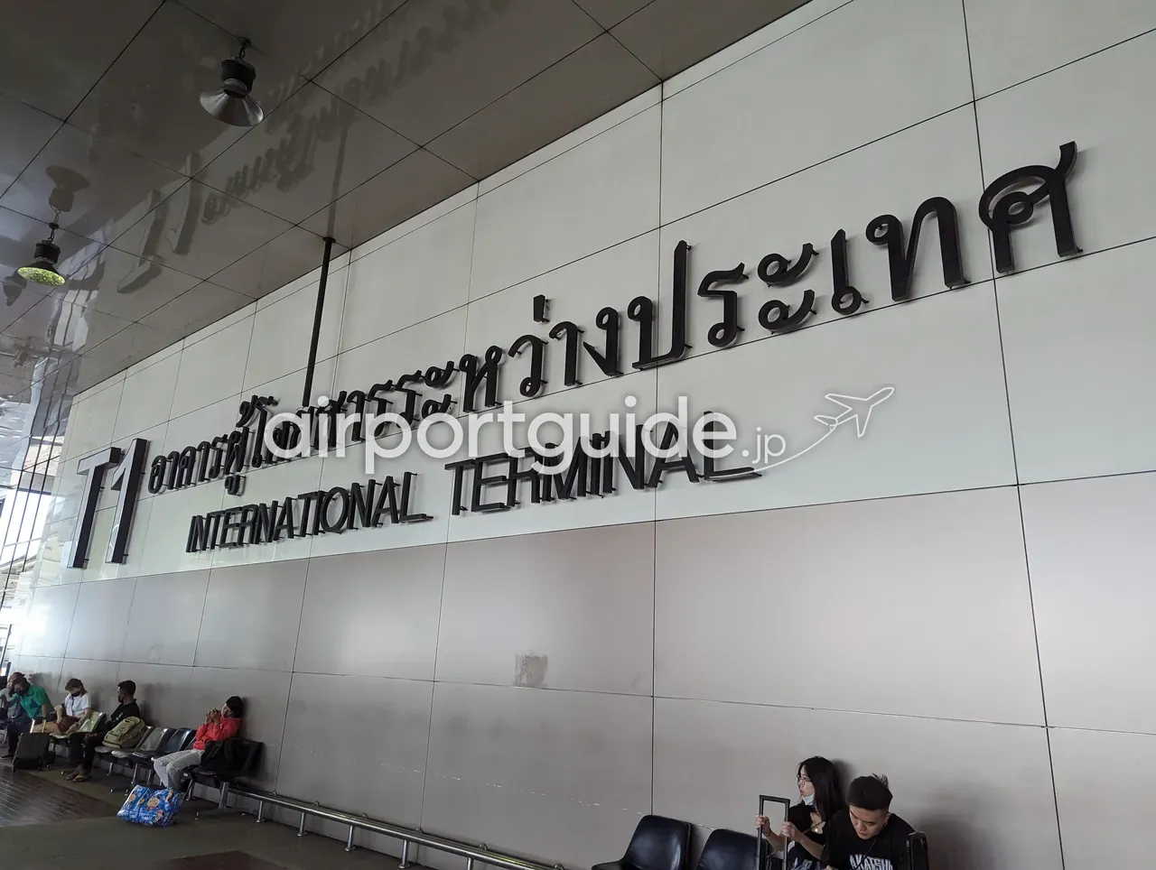 「T1 INTERNATIONAL TERMINAL」と大きく掲げられた壁面サイン。タイ語（อาคารผู้โดยสารระหว่างประเทศ）が併記され、下のベンチに旅客が座っている。