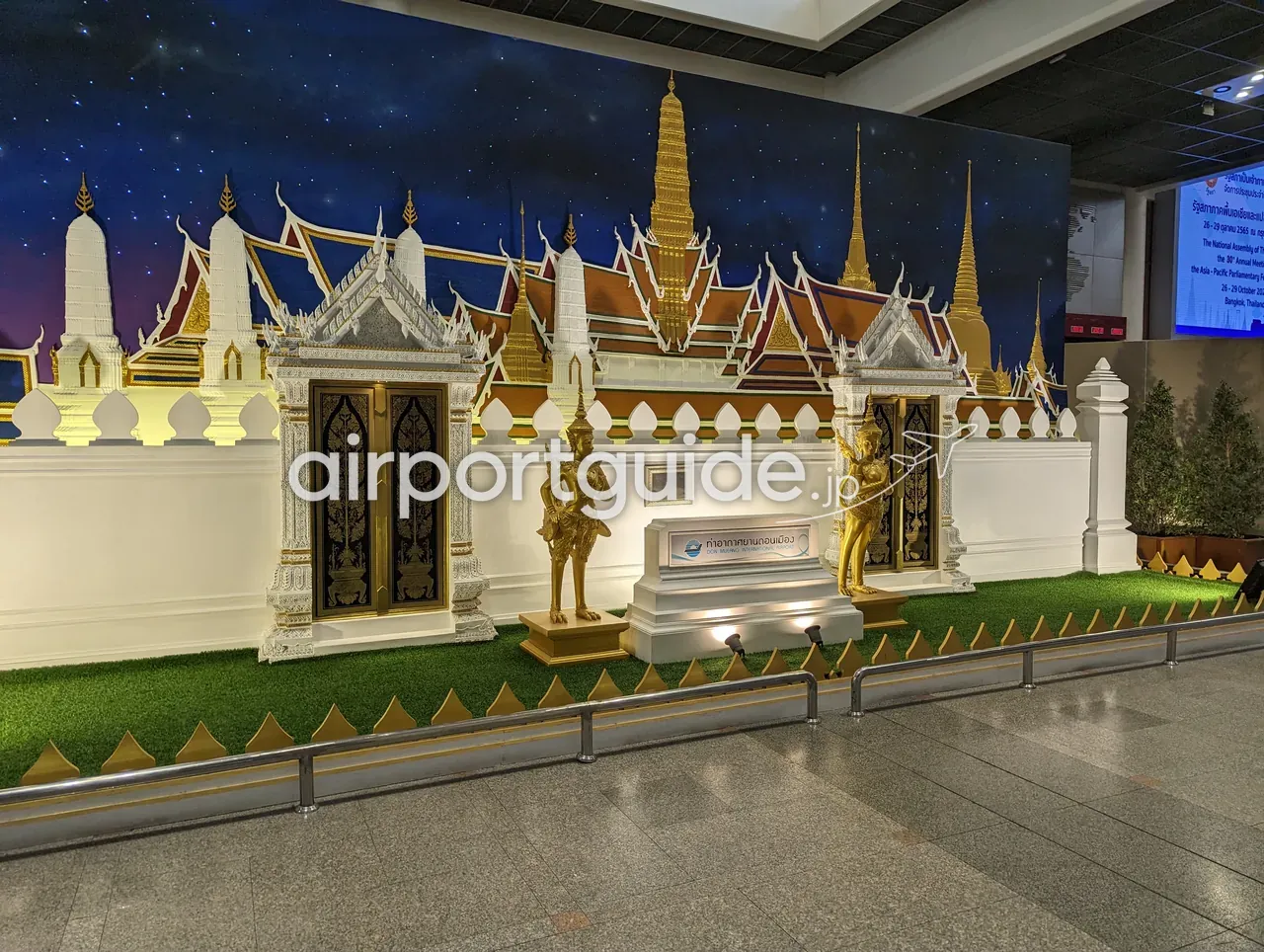 ドンムアン空港内のタイ王宮を模した装飾展示。白と金の宮殿風の壁と仏塔、黄金の守護神像（ヤック）、人工芝と金色の柵、中央に「ท่าอากาศยานดอนเมือง DON MUEANG INTERNATIONAL」の銘板が置かれる。