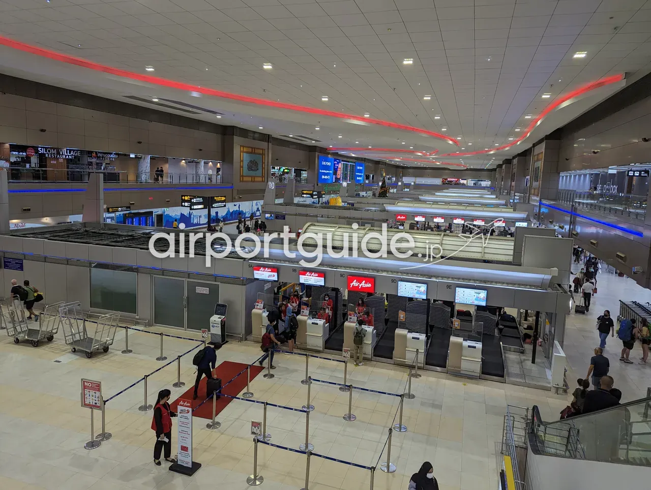 上階から見下ろしたドンムアン空港のチェックインホール。赤い「AirAsia」のカウンター（9A〜9E）に旅客が並び、天井には赤いラインの照明、奥に「SILOM VILLAGE RESTAURANT」の看板と手荷物カートが見える。