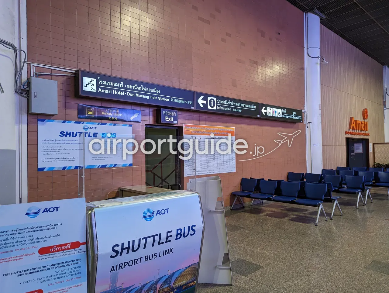 ドンムアン空港の待合エリアにあるAOTの「SHUTTLE BUS／AIRPORT BUS LINK」案内カウンター。頭上の案内サインに「Amari Hotel・Don Mueang Train Station」「Railway Station」への方向表示と時刻表、青い待合椅子が並ぶ。