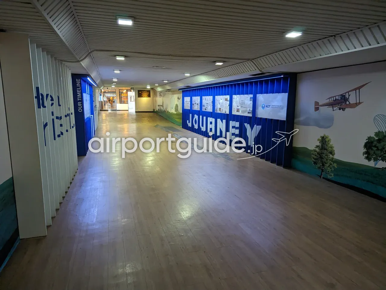ドンムアン空港の連絡通路に設けられた航空史の展示。「OUR TIMELINE」「JOURNEY」の青い装飾壁とパネル、右手の壁に緑の草原と初期の複葉機を描いた壁画、木目の床が続く。
