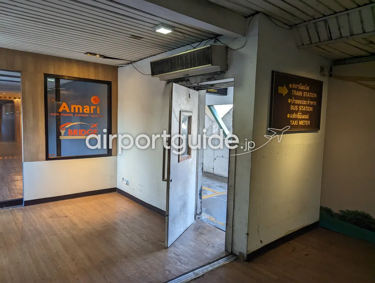 ドンムアン空港の連絡通路の入口。窓に「Amari DON MUANG AIRPORT · BANGKOK · THE BRIDGE」のロゴ、右の案内板に「TRAIN STATION →／BUS STATION／TAXI METER」の方向表示、奥に屋根付きの通路へ続くドアがある。