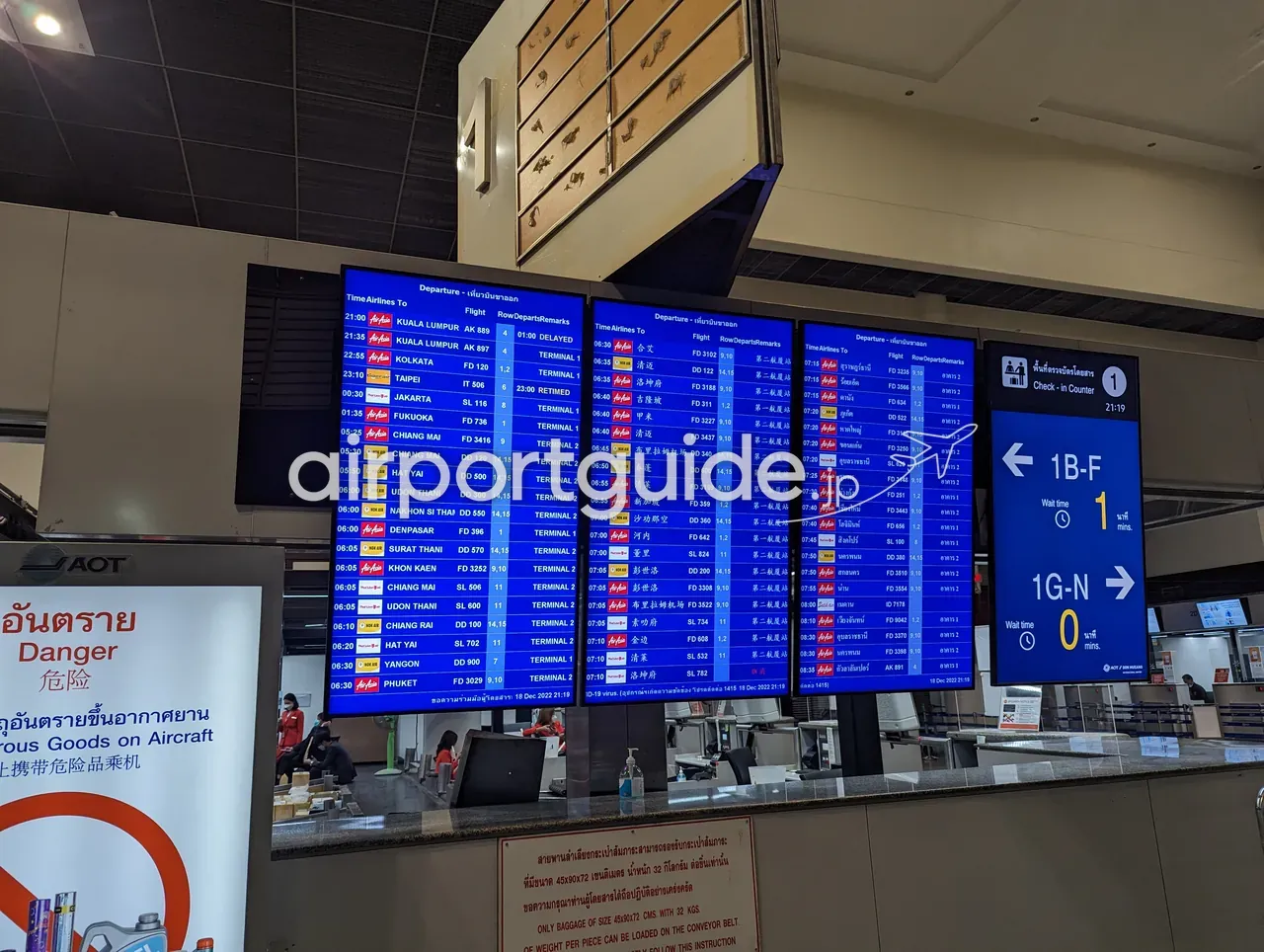 ドンムアン空港の青いLED出発案内板が3面並び、エアアジアやノックエアなどの便名・行き先（クアラルンプール、台北、福岡、チェンマイ、ハジャイほか）と「TERMINAL 1／TERMINAL 2」の別、右にチェックインカウンター1の案内が表示されている。