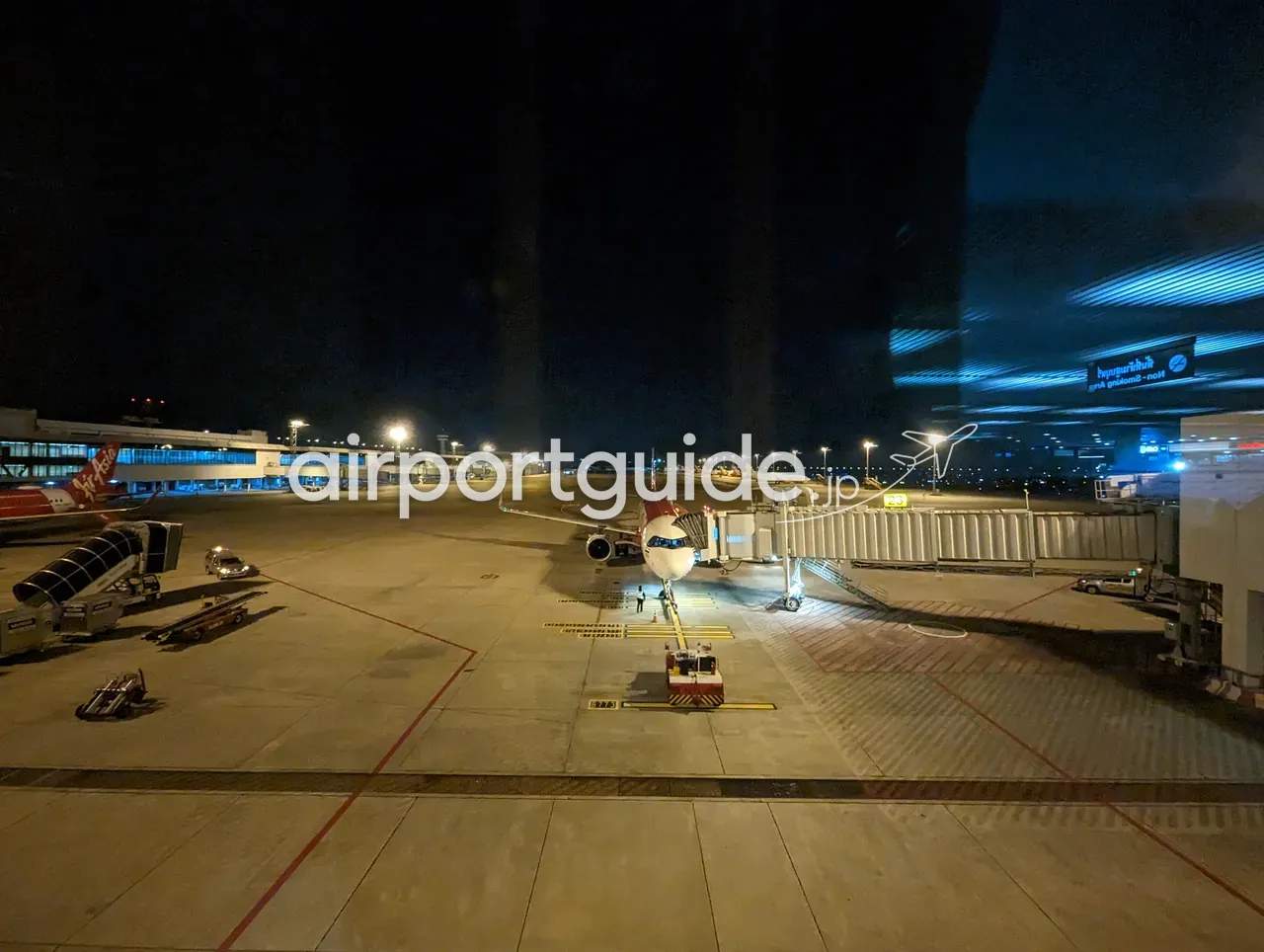 夜のドンムアン空港のエプロン。ボーディングブリッジに接続して駐機するエアアジアなどの機体、赤い尾翼、誘導路の灯りとターミナル建物、「Non-Smoking Area」のサインが見える。