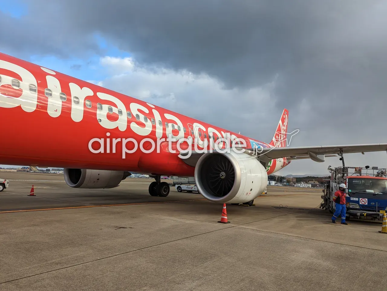 福岡空港のエプロンに駐機するエアアジア（airasia.com）の赤い機体を間近から見上げた構図。胴体の白いロゴとエンジン、翼下で作業する青い作業服の地上スタッフと給油車。