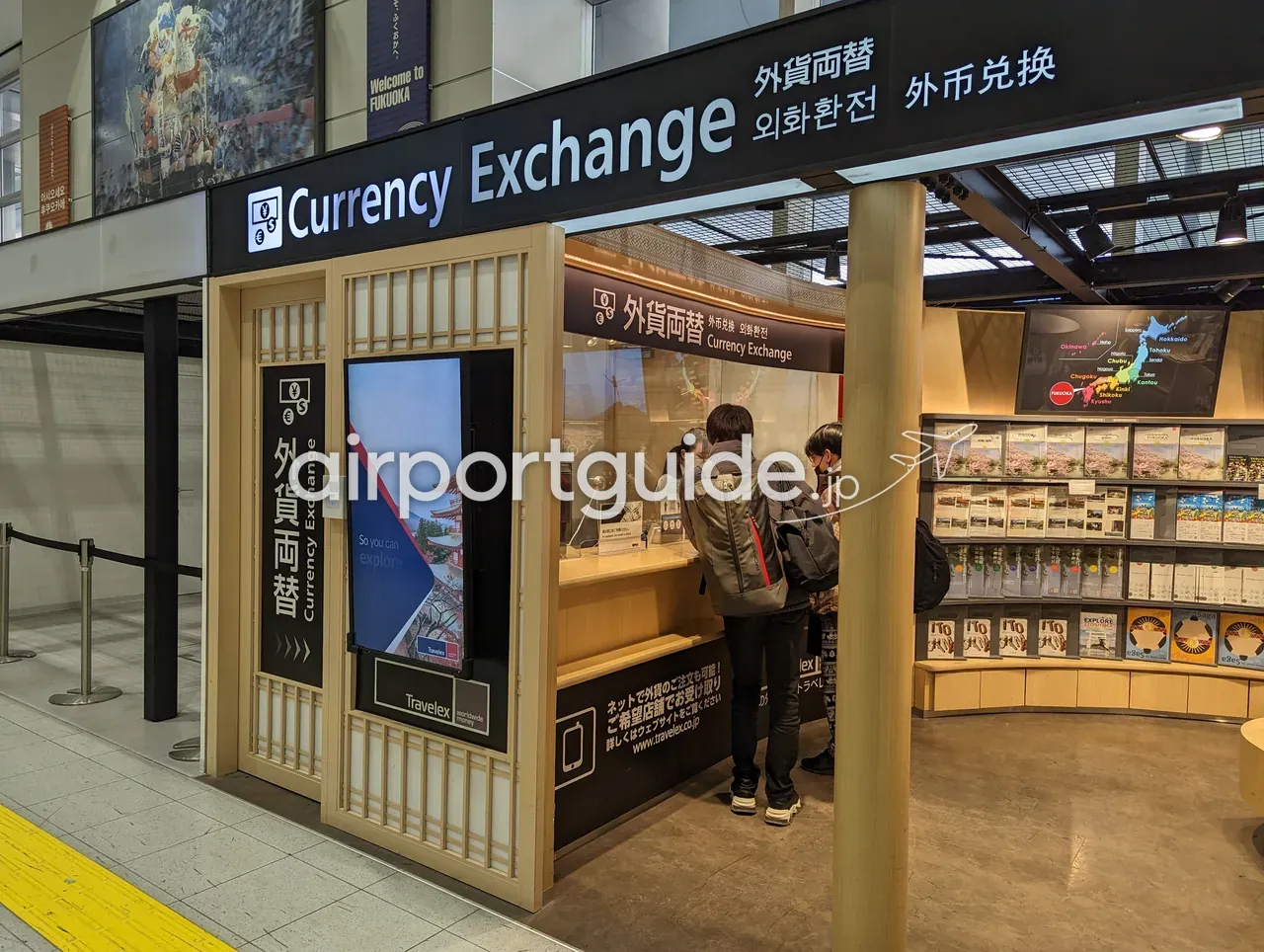 福岡空港のトラベレックス（Travelex）で外貨両替をする利用客。「Welcome to FUKUOKA」のバナーと九州の観光パンフレット棚が並ぶ。