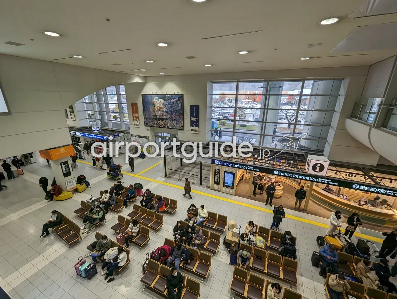 福岡空港国際線ターミナルの到着ロビーを上階から見下ろしたカット。待合いすに座る多数の到着客、「Currency Exchange 外貨両替」と「総合案内所 Tourist Information」の窓口、バス券売り場、壁面に博多祇園山笠の飾り山の写真パネル。