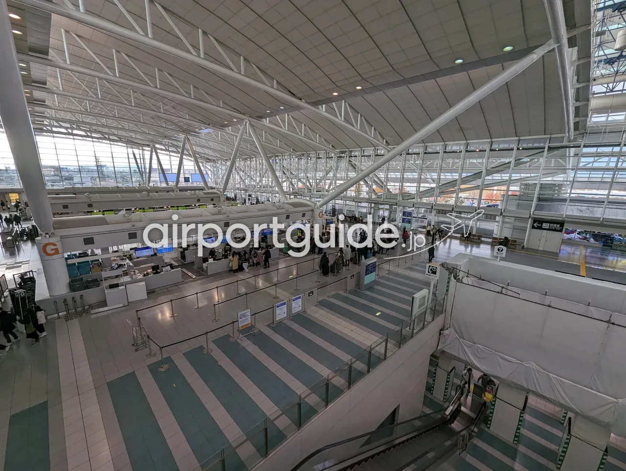 福岡空港の出発ロビー全景を上階から俯瞰。白いテント状のトラス屋根の下に「G」の柱とチェックインカウンターが並び、緑・白のストライプ床、下階へ続くエスカレーター。
