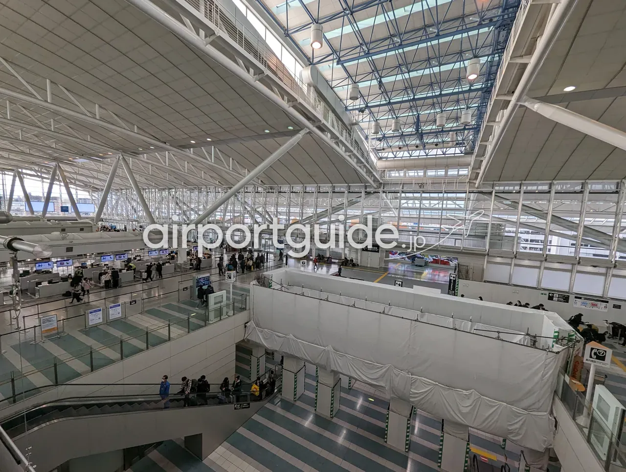福岡空港ターミナルの吹き抜けの大空間を上から見下ろした広角。青く塗られた鉄骨のトラス天井と、下階へ続くエスカレーター、白い養生シートに覆われた連絡通路が見える。