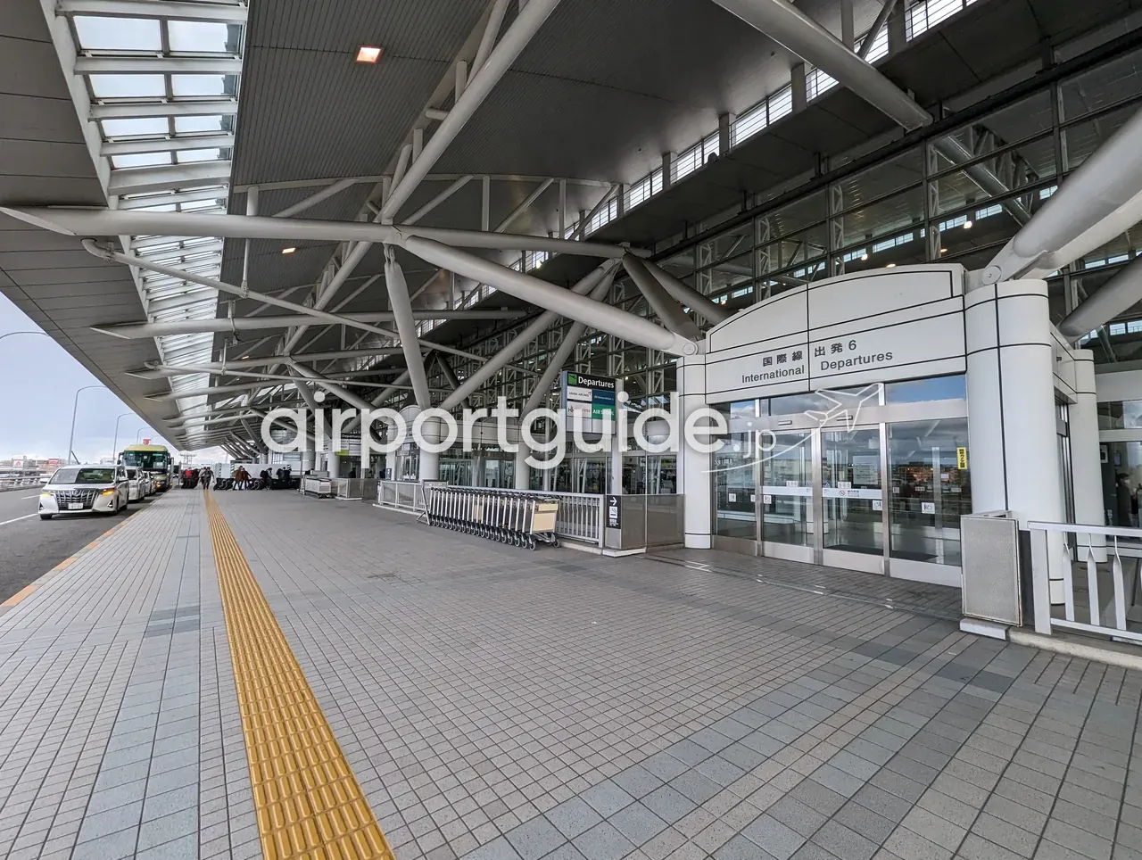 福岡空港国際線ターミナルの出発階カーブサイド。「国際線 出発6 International Departures」の入口サインと自動ドア、手荷物カート置き場、左手の車道にタクシーやバスが並ぶ。