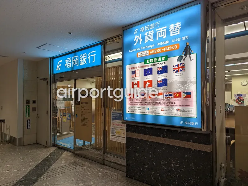 福岡銀行 外貨両替（Currency Exchange）。取扱通貨の国旗を並べた案内ボードと、福岡銀行 福岡空港支店の入口。両替機と有人カウンターがある。