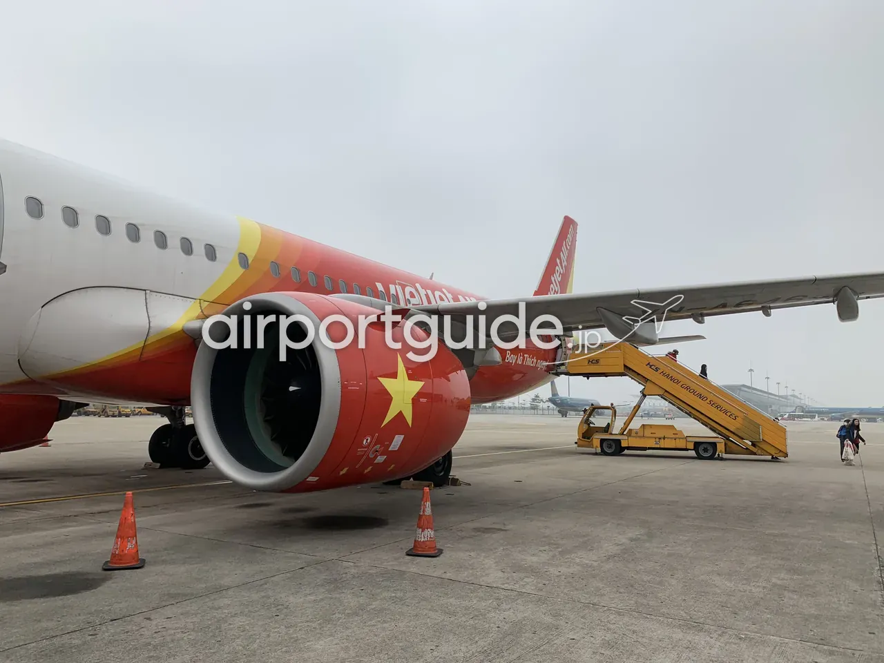ノイバイ空港の駐機場で、ベトジェットエアのA320のエンジン（赤い覆いに黄色い星、「Bay là Thích ngay」）とタラップを間近に。「HGS Hanoi Ground Services」のボーディングステップと、歩いて搭乗する乗客、オレンジのコーン。
