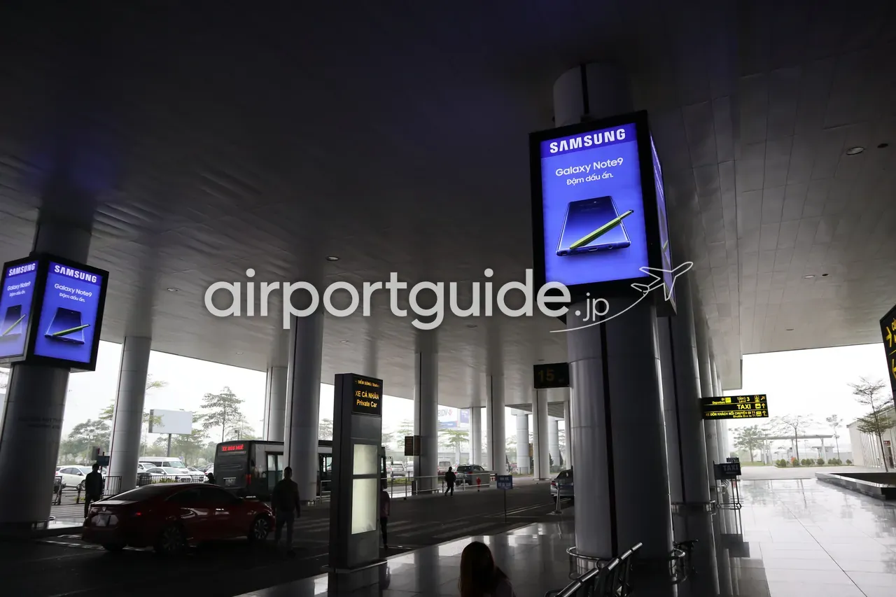ノイバイ空港ターミナルの屋根付き車寄せ（カーブサイド）。柱にSamsung Galaxy Note9のデジタル広告、「XE CÁ NHÂN / Private Car（自家用車）」のブースと「TẦNG 3 Departures / TAXI」の黄色い案内サイン、赤い乗用車が停まる。