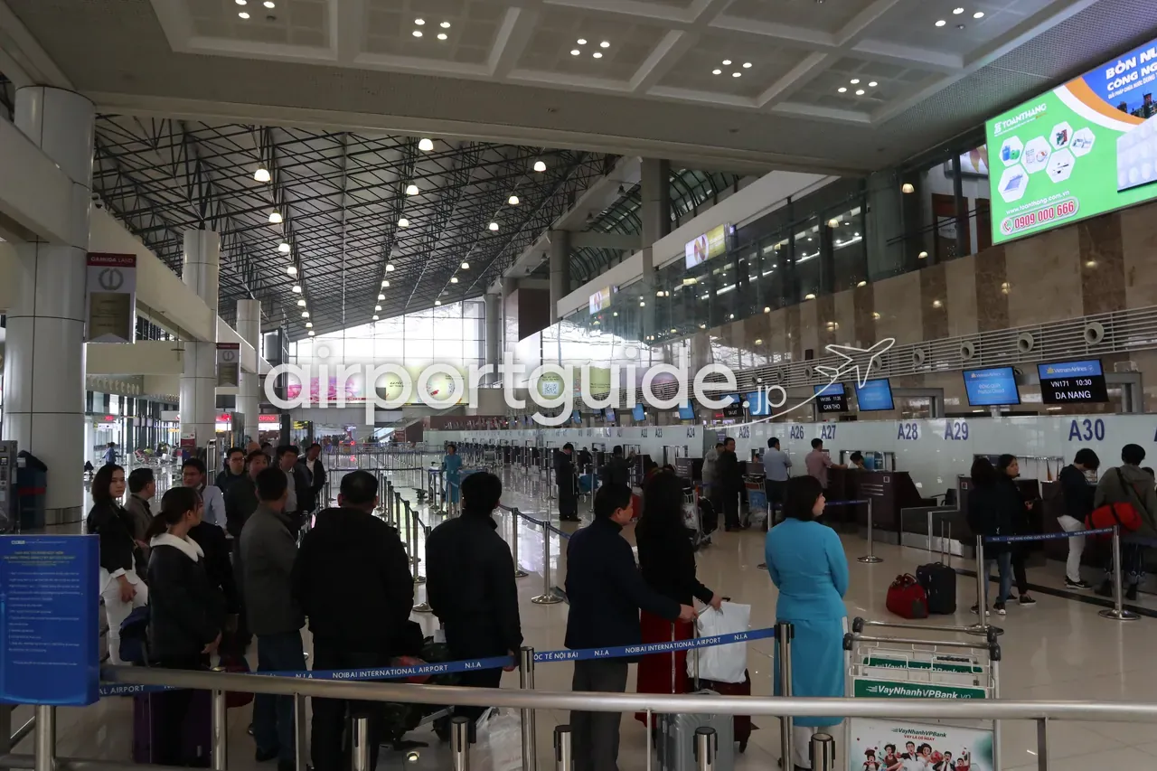 ノイバイ空港・国内線のチェックインエリア（A23〜A30付近）。「VN171 10:30 DA NANG」のフライト表示、アオザイ姿のベトナム航空スタッフ、規制ベルトに「NOIBAI INTERNATIONAL AIRPORT N.I.A」の印字、行列する旅客。