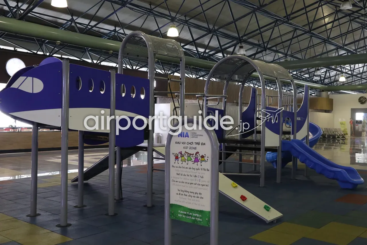 ノイバイ空港の子ども遊び場。「NIA NOIBAI INTERNATIONAL AIRPORT / KHU VUI CHƠI TRẺ EM / KID ZONE」の看板、青い飛行機型の大型遊具とすべり台、「PLEASE PLAY SAFELY! / HÃY VUI CHƠI AN TOÀN!」の注意書き。