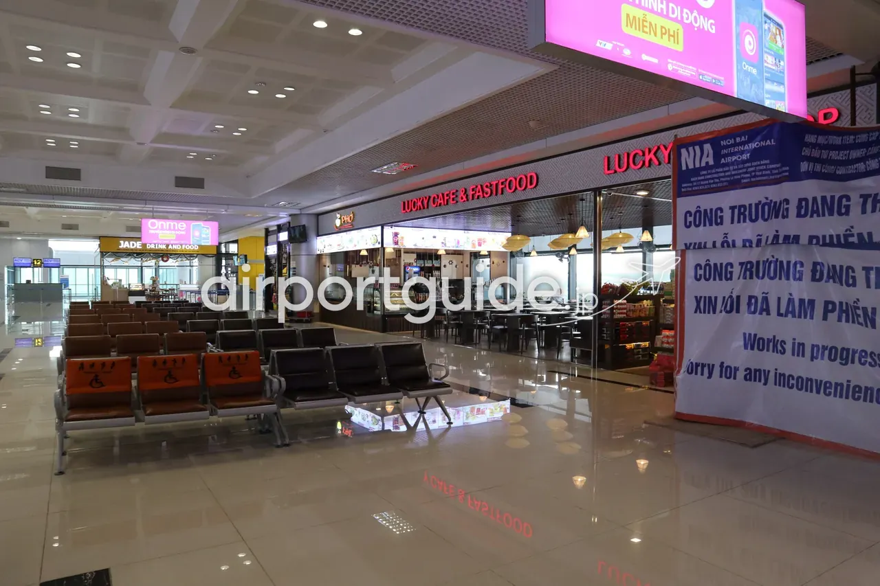ノイバイ空港・国内線出発エリアの待合座席と飲食店。「LUCKY CAFE & FASTFOOD」「JADE DRINK AND FOOD」「PHỞ」の店、連結ベンチ（一部は優先席）、ピンクのアプリ広告、工事中の仕切り「Works in progress」。